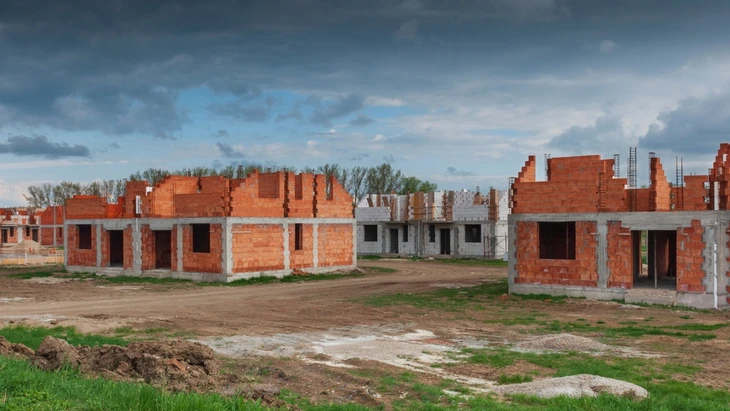 Photographie d'un chantier de construction avec une équipe de trois ou quatre ouvriers professionnels travaillant en coordination : au premier plan, un ouvrier pose ou finit une dalle béton avec un outil de lissage, un deuxième ouvrier prépare le terrain ou supervise la mise en place du coffrage, un troisième gère les matériaux ou le drainage. Arrière-plan montrant un terrain préparé pour construction avec bâtiment agricole ou zone industrielle visible. Éclairage naturel, équipes portant équipement de sécurité professionnel. La scène doit clairement montrer la coordination entre plusieurs corps de métier travaillant ensemble.